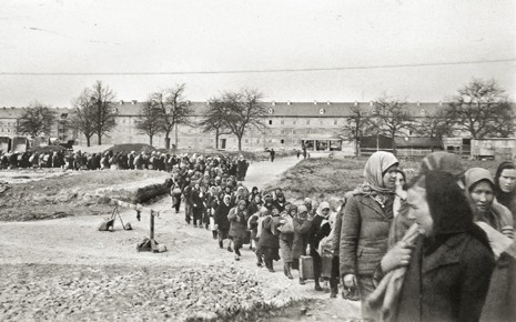Historische Ansicht in schwarz weiß. Eine große an Menschen zieht sich durch das Bild von rechts vorne bis weit links hinten. Vorne sieht man einige Frauen mit Kopftuch im Profil. Im Hintergrund sieht man ein langes Gebäude mit zwei Stockwerken und einige kahlen Bäume.