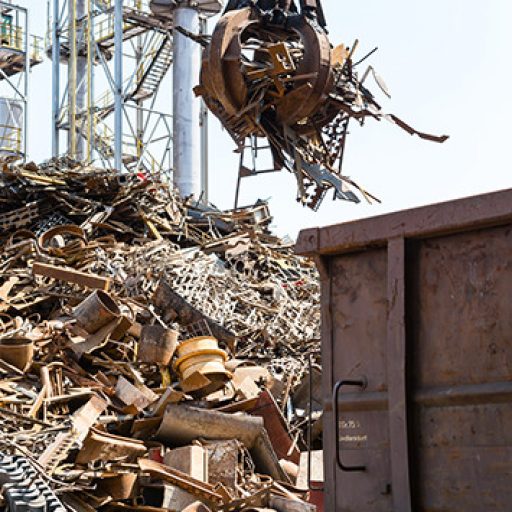 Industrieller Greifarm hebt Metallschrott auf einen großen Schrotthaufen vor einer Industrieanlage, daneben ein Güterwaggon.