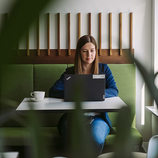 Bianca sitzt mit einem Kaffee und Laptop an einem Tisch und arbeitet.