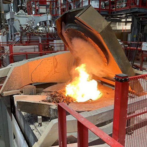 Industrieanlage zur Schrottverarbeitung mit Schmelzofen, in dem Metallschrott erhitzt wird. Flammen und glühendes Material sichtbar, umgeben von Sicherheitsvorrichtungen in einer Produktionshalle.