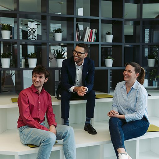 Justin is sitting together with 2 other voestalpine employees in a modern environment and they are all looking at something outside the picture to their right.