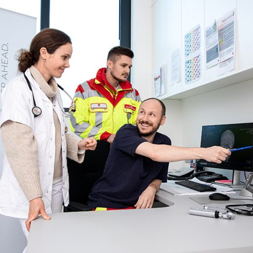 Zwei Männer und eine Frau schauen auf zwei Bildschirme und sprechen sich über medizinische Themen ab.