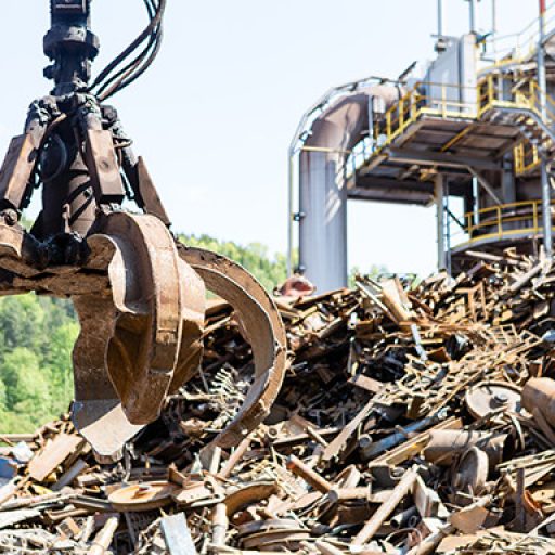 Industriekran mit Greifarm hebt Metallschrott auf einem großen Schrotthaufen vor einer Industrieanlage.