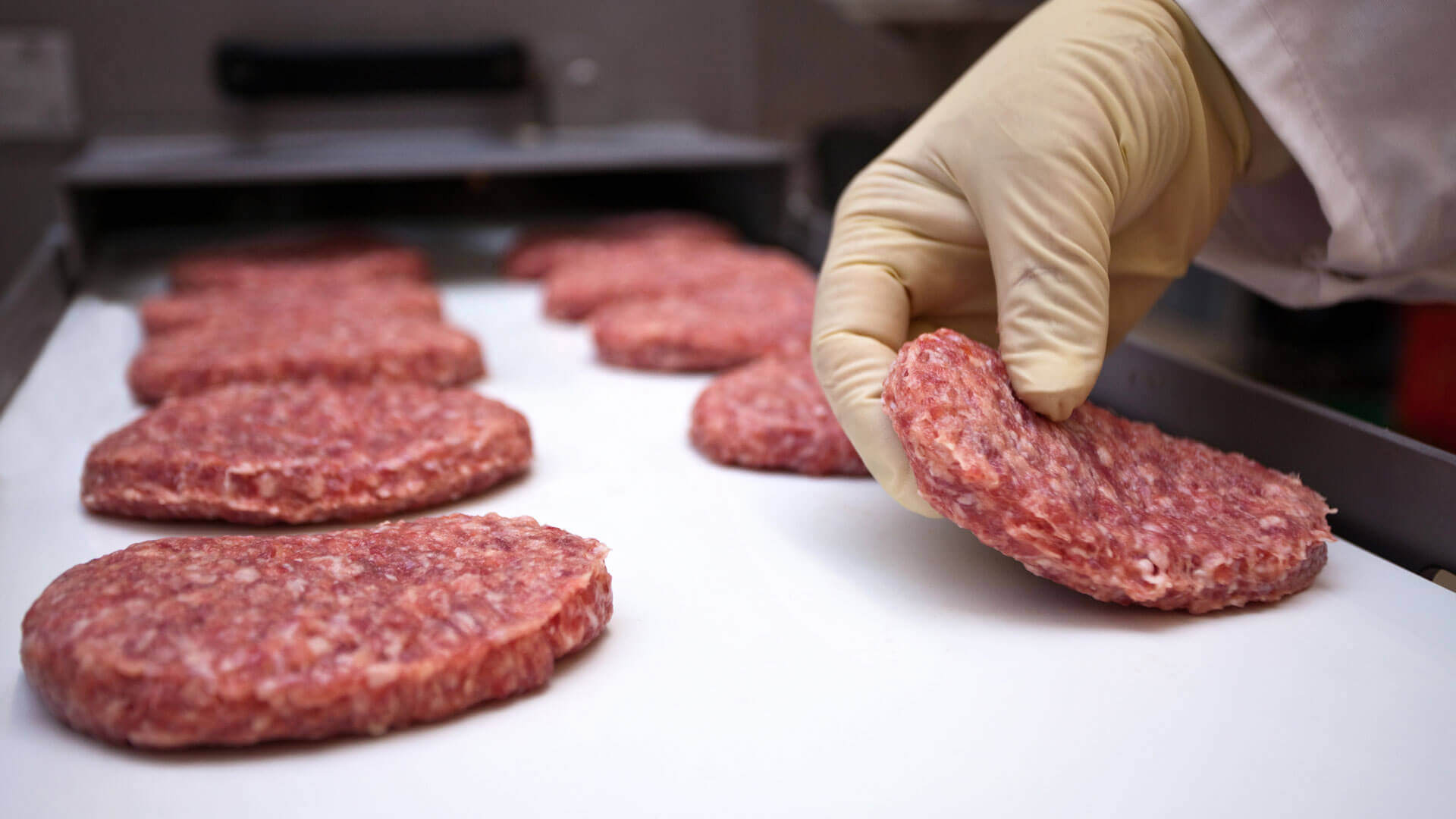 A gloved hand picks up a meat pattty on a production line.