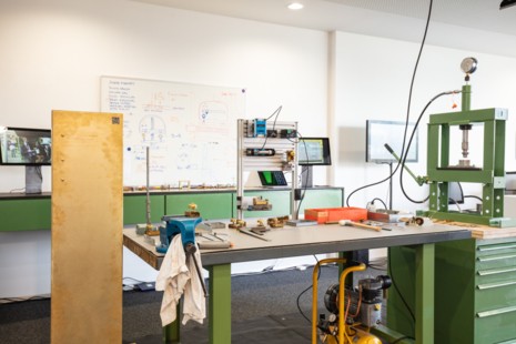 Eine Werkbank im Vordergrund, auf der Werkzeug liegt. Daneben eine messingfarbene Stele. Dahinter eine Wand, auf der Skizzen gezeichnet sind